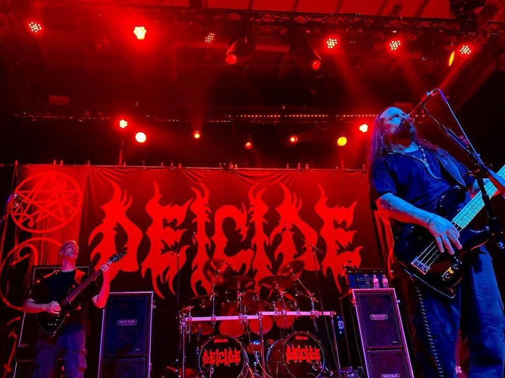 Dos integrantes de Deicide tocando en vivo en un concierto, con una batería en segundo plano y un ambiente iluminado en tonos rojos