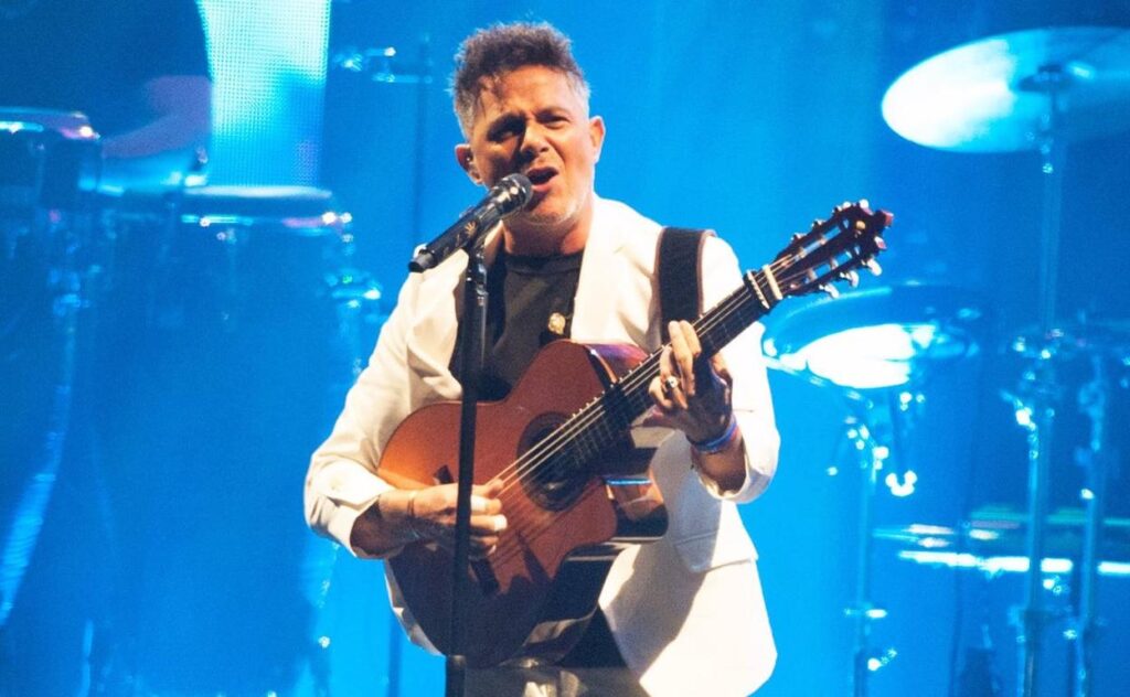 Alejandro Sanz toca su guitarra en un escenario iluminado por luces azules, luciendo una elegante ropa en blanco y negro