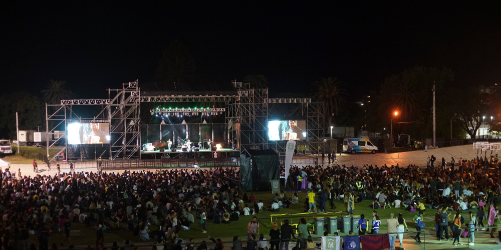 Noche de concierto al aire libre en el Velódromo Municipal de Montevideo, con una multitud de espectadores disfrutando del evento musical sentados en el césped y de pie, iluminados por la luz del escenario y pantallas gigantes