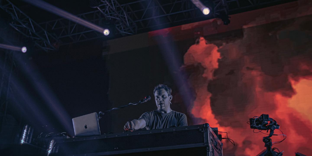 Juan Hansen vestido con camisa negra, con un fondo de humo rojo y con su laptop y micrófono