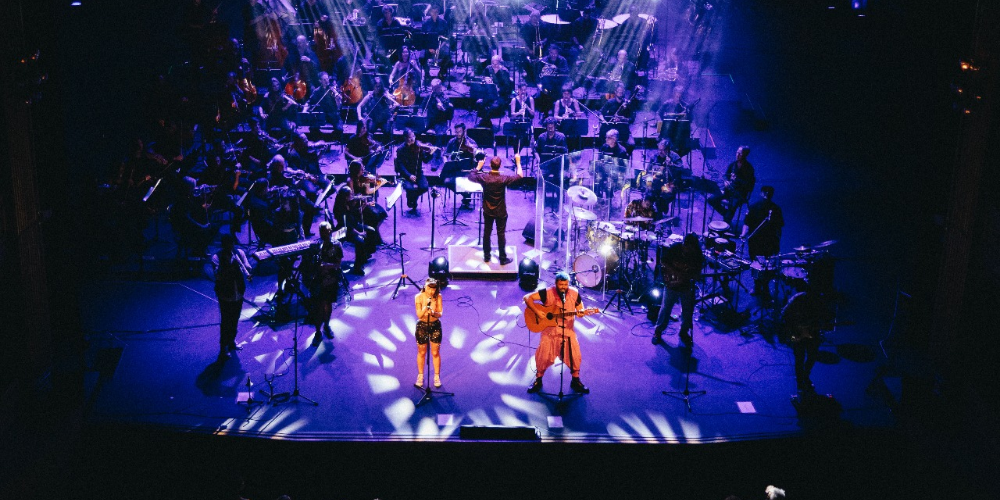 Los cantantes de Cuatro Pesos de Propina en el escenario, mientras un director guía a los músicos en segundo plano