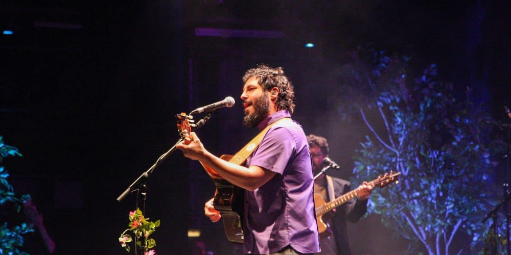 El Kanka cantando en vivo en un escenario, llevando una camisa violeta y una guitarra cruzada