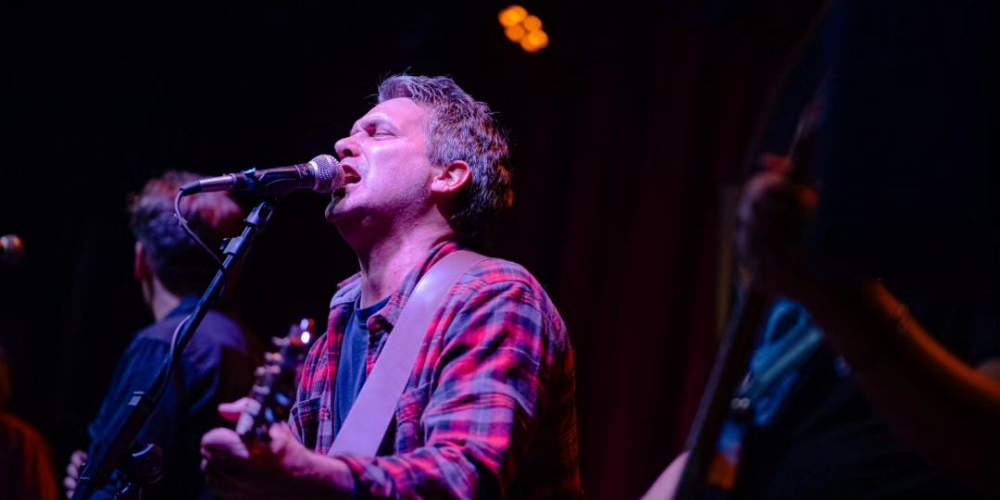 Rodrigo Manigot, vocalista de Ella Es Tan Cargosa, cantando con intensidad con los ojos cerrados y vistiendo una camisa a cuadros