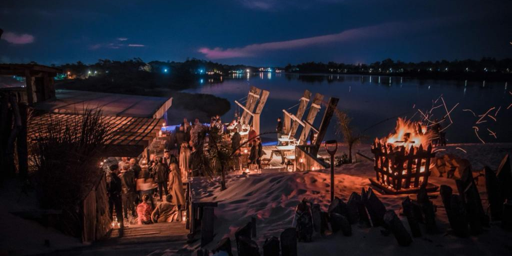 Vista aérea nocturna de Burdeos Restaurant, destacando una fogata encendida y un cálido grupo de personas disfrutando, todo enmarcado por la íntima iluminación y la naturaleza circundante en Ciudad de la Costa