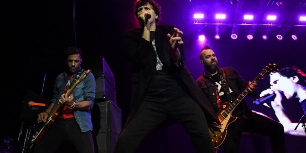 Andrés Ciro Martínez, vestido de negro, cantando apasionadamente en el micrófono, con dos músicos de Ciro y Los Persas tocando detrás de él en el escenario