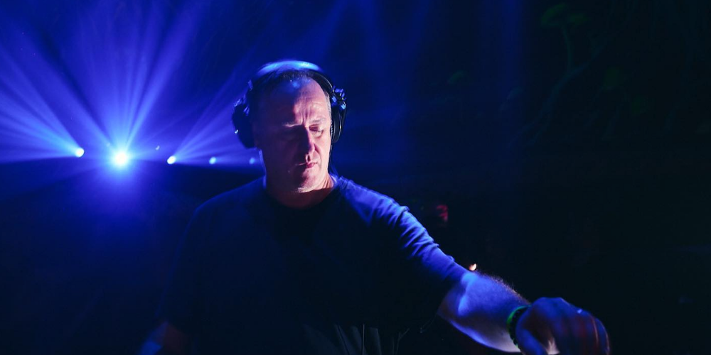 Sasha mezclando música en una cabina de DJ, con luces azules brillantes que irradian desde el fondo del escenario